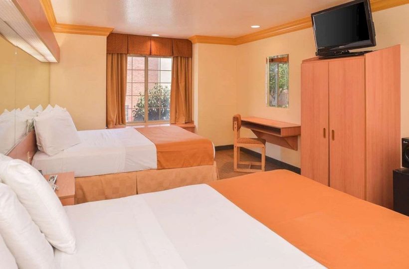 The image shows a hotel room with two beds, a window, a wardrobe, a wall-mounted TV, and a desk with a chair, all in warm tones.