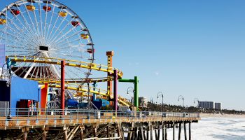 The image shows a coastal amusement park with a large Ferris wheel and roller coaster on a pier overlooking the ocean under a clear blue sky.