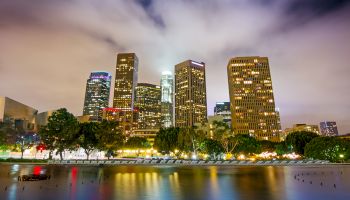 A cityscape image at night features illuminated skyscrapers, a calm water body with reflections, and trees in the foreground, under a cloudy sky.