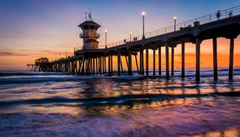 A pier extends into the ocean at sunset, lit up by lights, with waves crashing gently below and a colorful sky in the background.