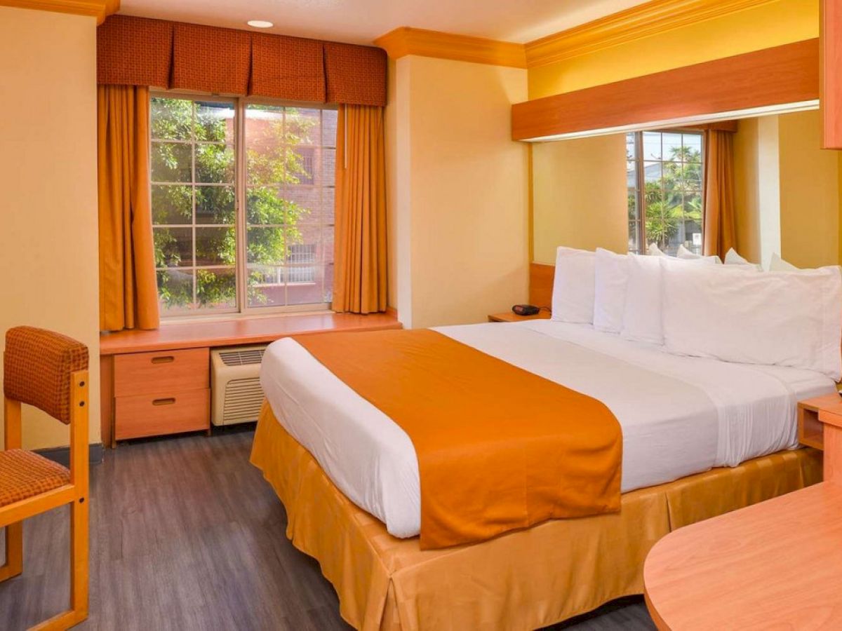 This image shows a cozy hotel room with a neatly made bed, a large window with curtains, a desk, and warm yellow and orange decor.