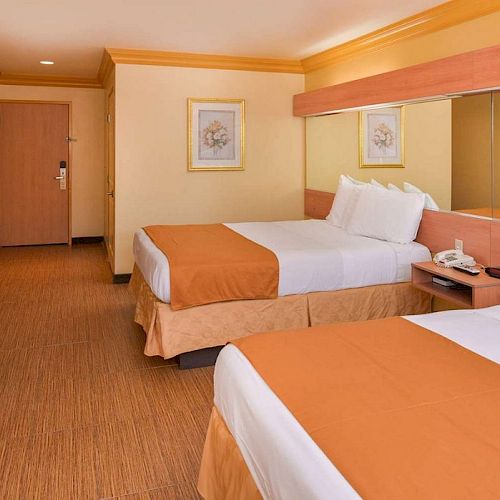 The image shows a hotel room with two double beds, a nightstand, wall art, a TV, and a door in the background.