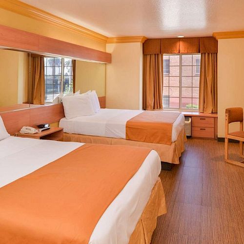 The image shows a hotel room with two double beds, orange and white bedding, a window, desk with chair, wall-mounted TV, and wooden furnishings.