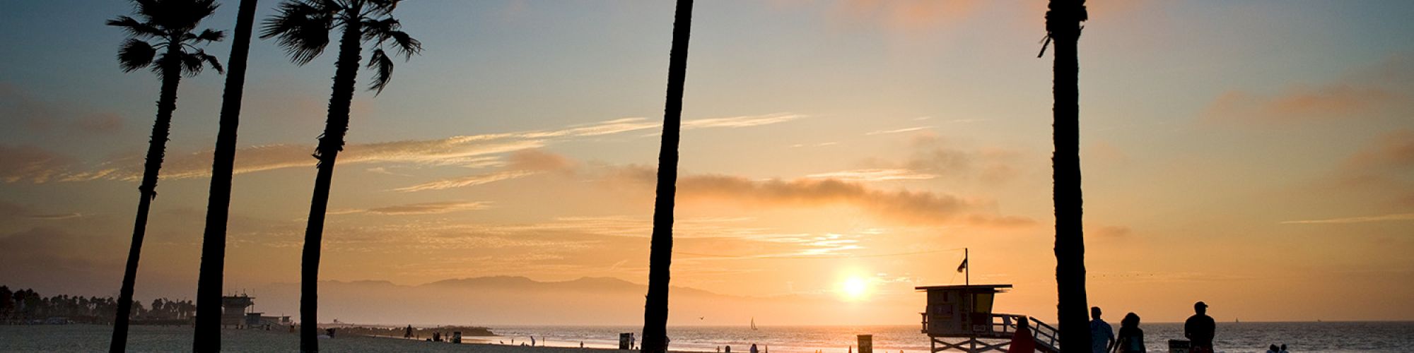 A beach at sunset with silhouetted palm trees, a lifeguard tower, and people walking in the sand.