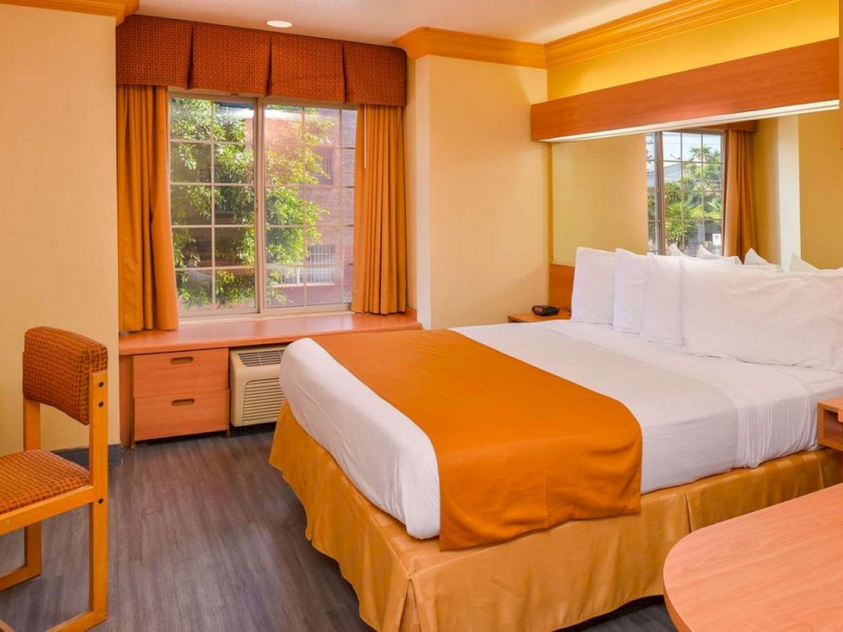 A clean hotel room features a queen-sized bed with orange bedding and pillows, a desk with a chair, and a window with a view.