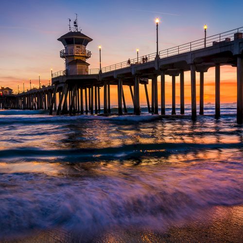 A pier extends over the ocean at sunset with people walking and streetlights illuminating the scene, while waves gently crash on the shore.