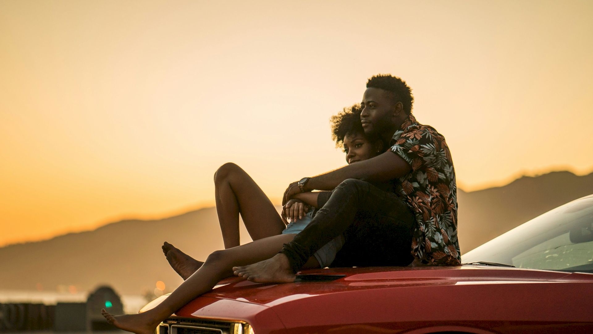 A couple is sitting on the hood of a red car, embracing and enjoying a sunset by a coastal road with mountains in the background.