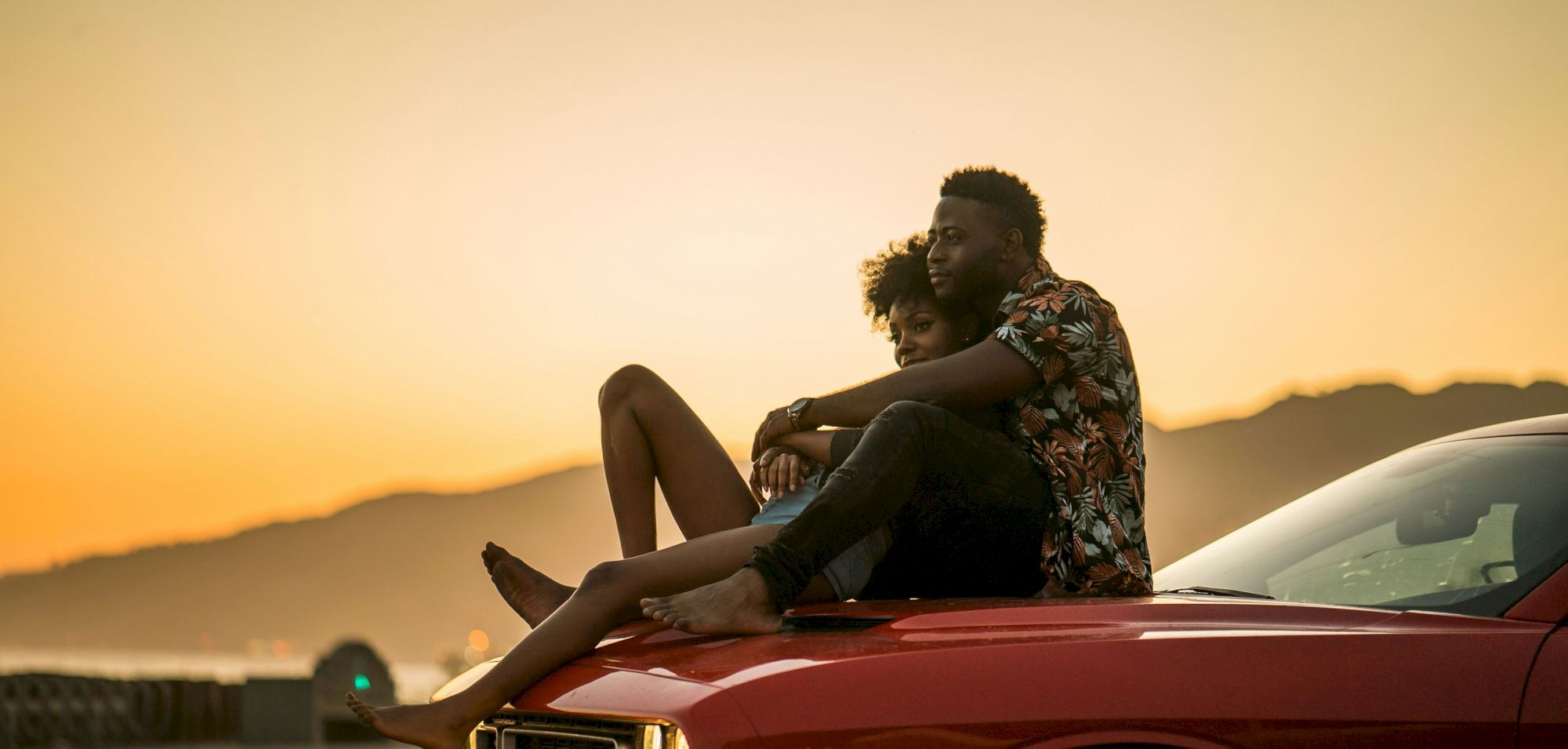 A couple is sitting on the hood of a red car, embracing and enjoying a sunset by a coastal road with mountains in the background.