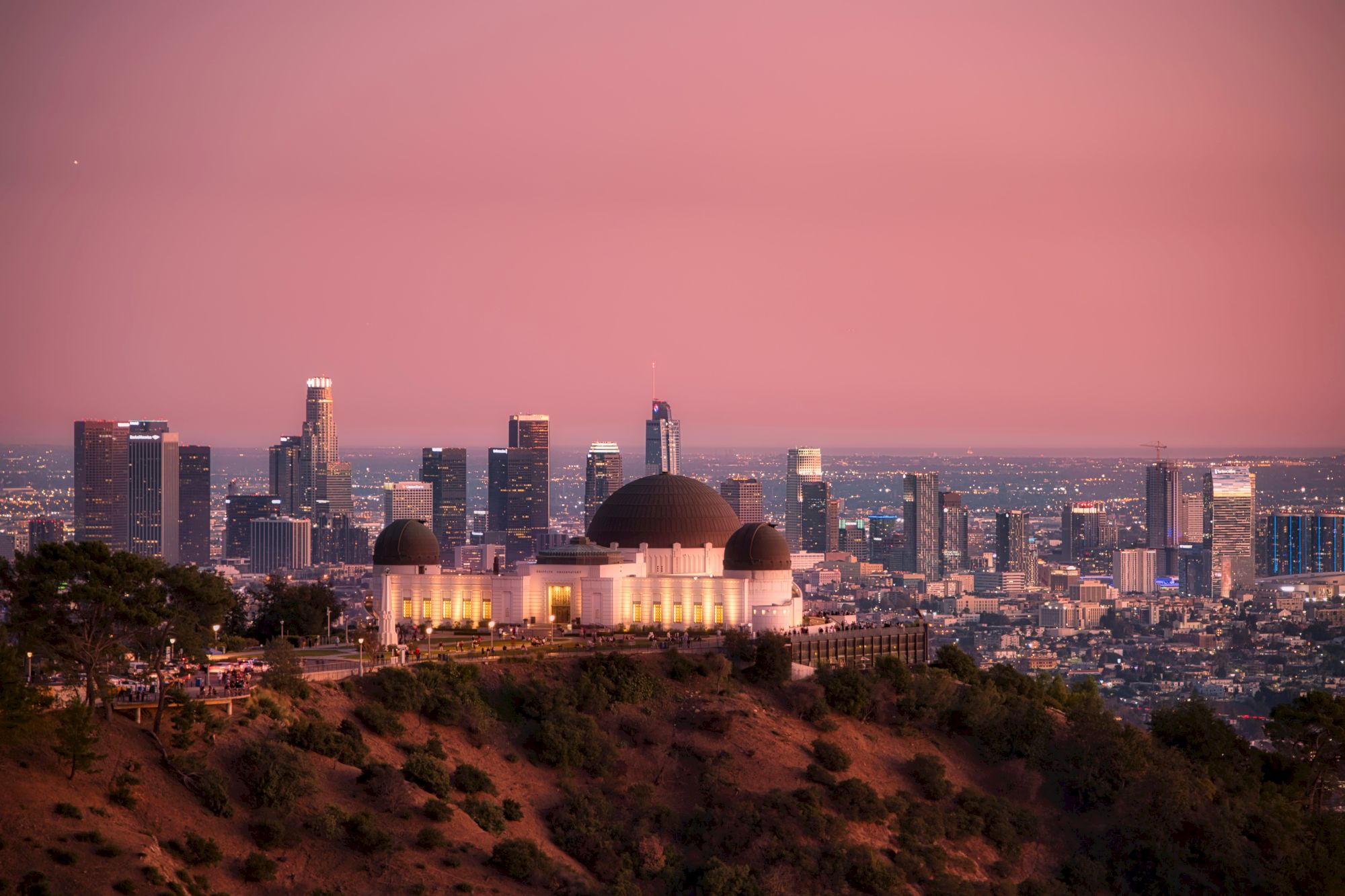 An observatory sits on a hill with a city skyline in the background under a pinkish sky at sunset. The cityscape shows various tall buildings.